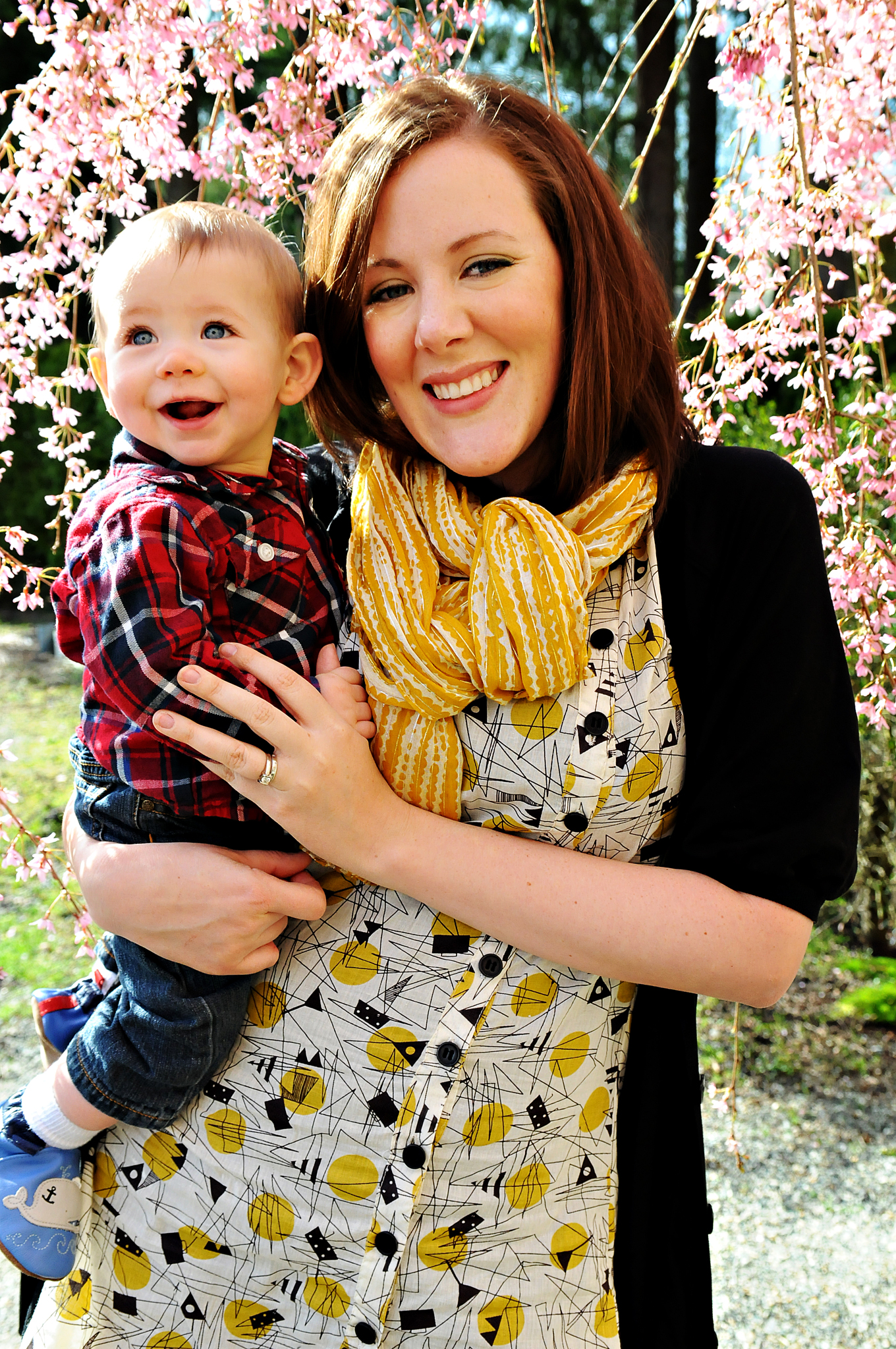 Mom holding her infant son while standing under a tree full of cherry blossoms. The boy wears plad and the mom wears a spring yellow scarf and dress.